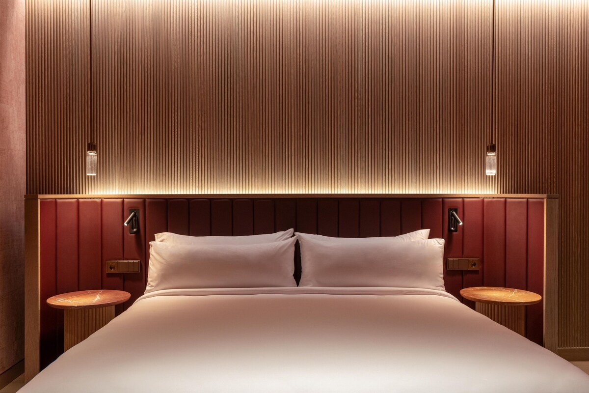 A hotel room with a large bed, two bedside tables, a wooden headboard, and wall-mounted lighting.