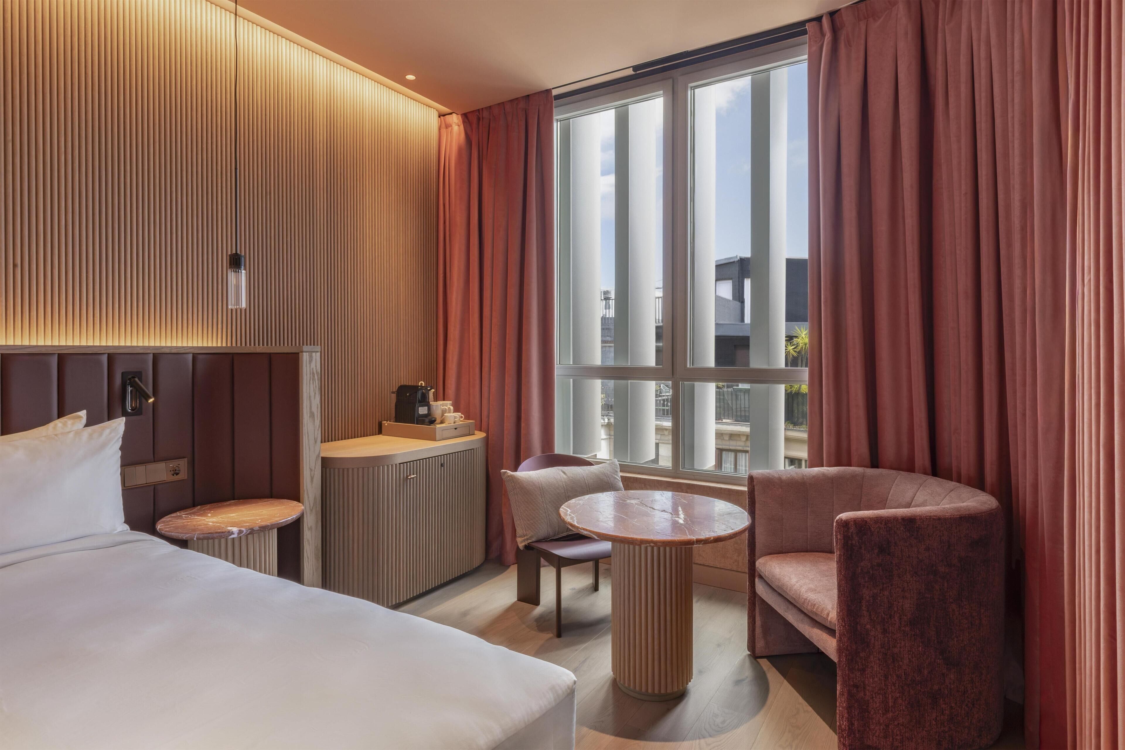 Habitación de hotel con cama, escritorio, silla, una mesa redonda y un ventanal con cortinas.
