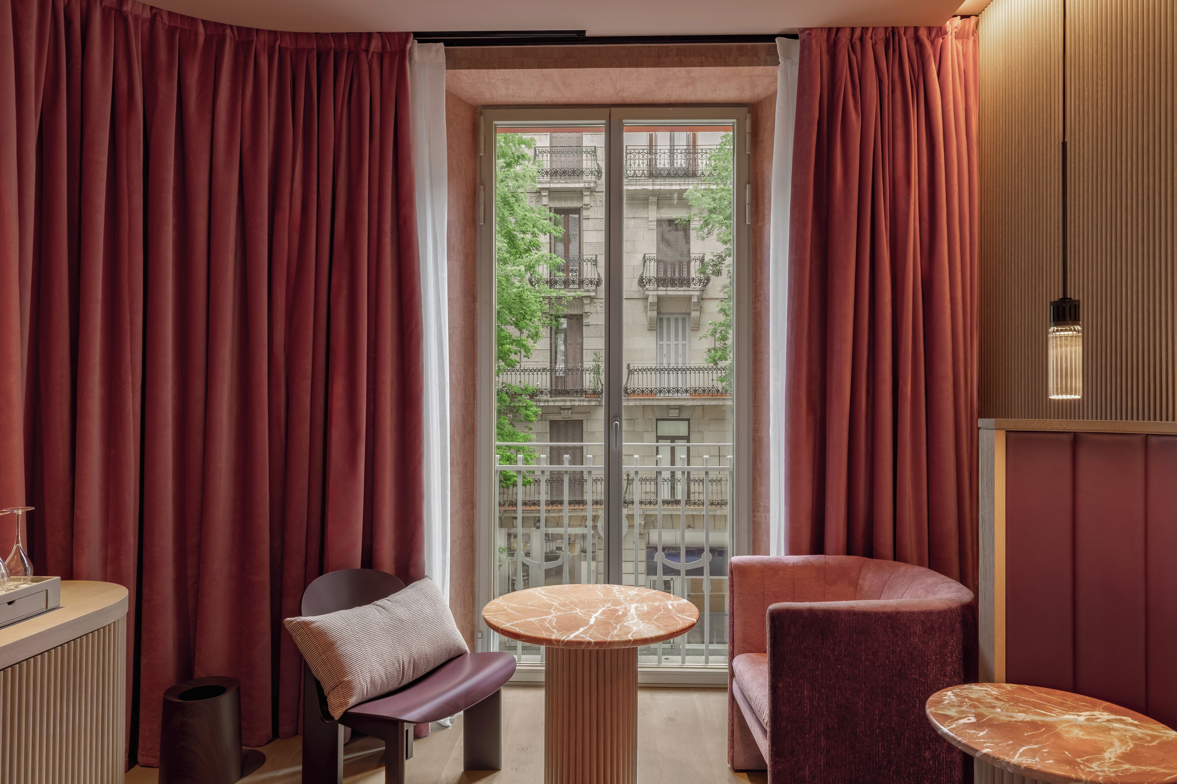 Une chambre d’hôtel avec un balcon, une table ronde, un fauteuil rembourré et un petit meuble.