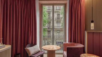 Une chambre d’hôtel avec un balcon, une table ronde, un fauteuil rembourré et un petit meuble.