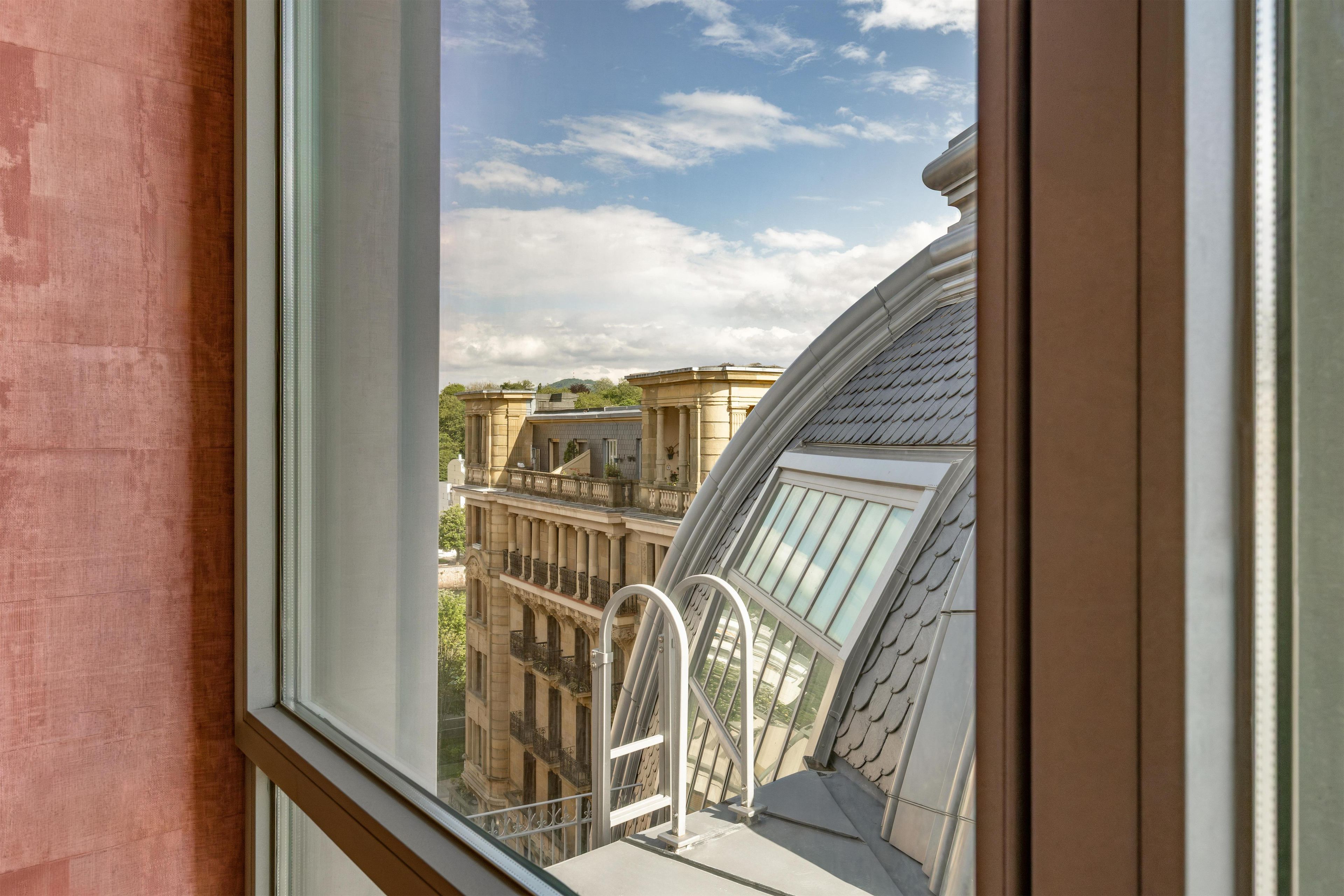 A view from a window with a balcony, overlooking a building with classical architecture and a clear sky.
