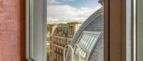 A view from a window with a balcony, overlooking a building with classical architecture and a clear sky.