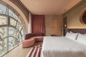 A modern hotel room with a curved seating area, a large bed, a round mirror, and a view of buildings through large windows. - Hotel Palacio Bellas Artes San Sebastián, Curio Collection (San Sebastian)