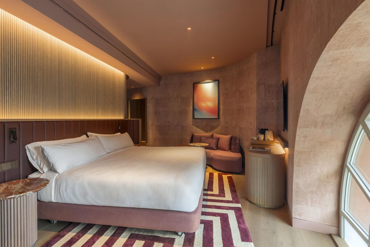 A modern hotel room with a large bed, a seating area, a TV, and a window.