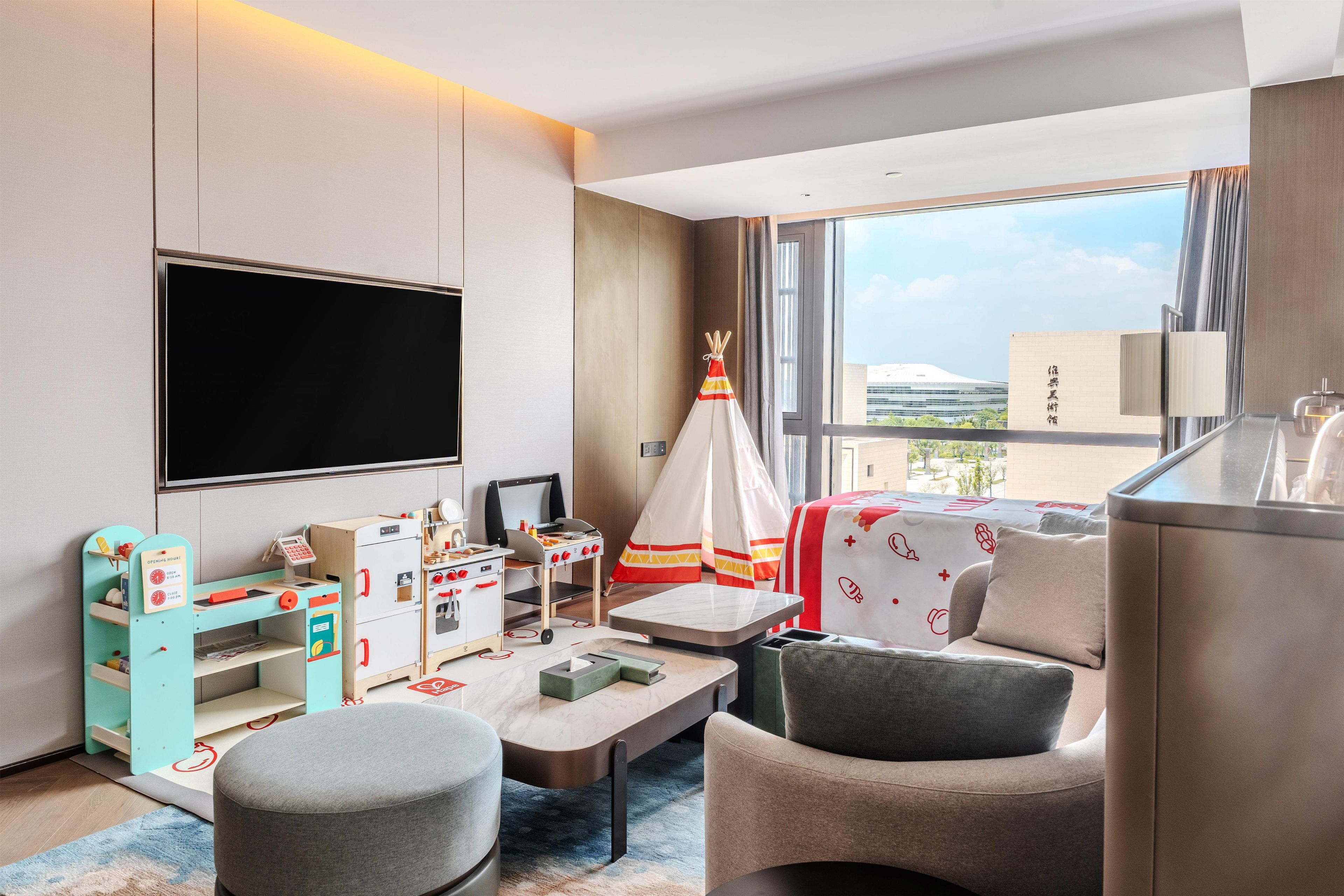 A modern hotel room with a large TV, a kitchenette, a comfortable seating area, and a view of the cityscape.