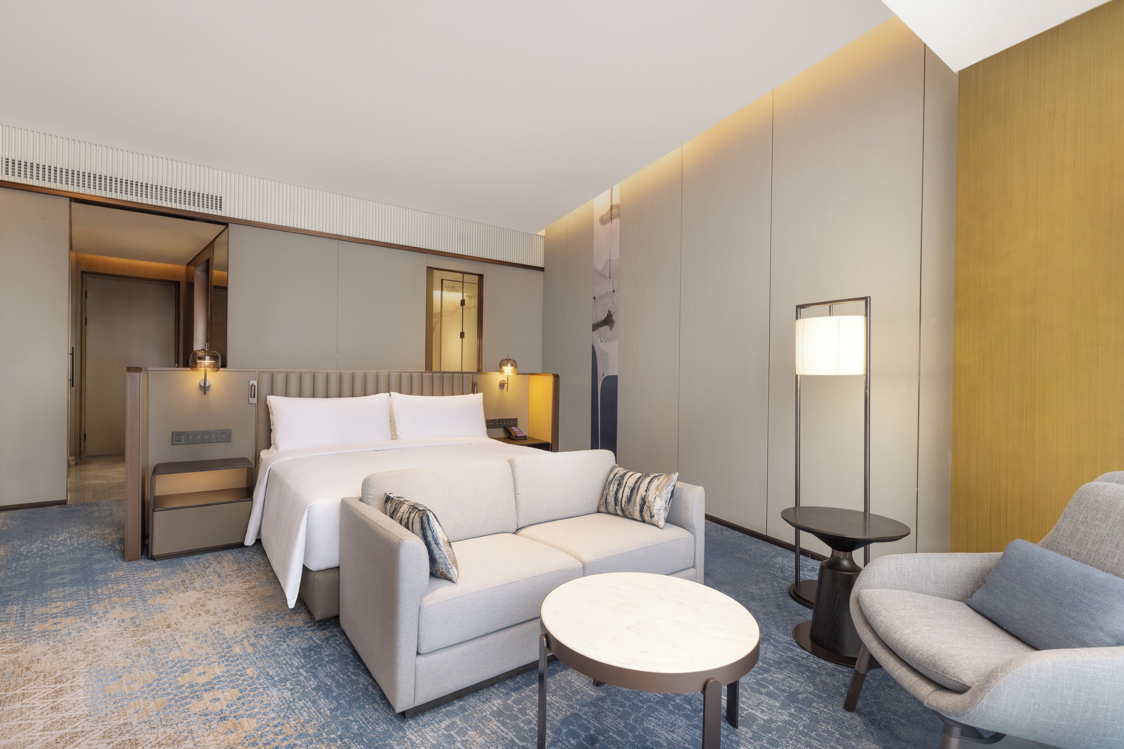 Habitación de hotel con cama, sofá, una mesita redonda y una silla. Se observa un buró con una lámpara y un teléfono. El piso es de madera y hay un ventanal grande.
