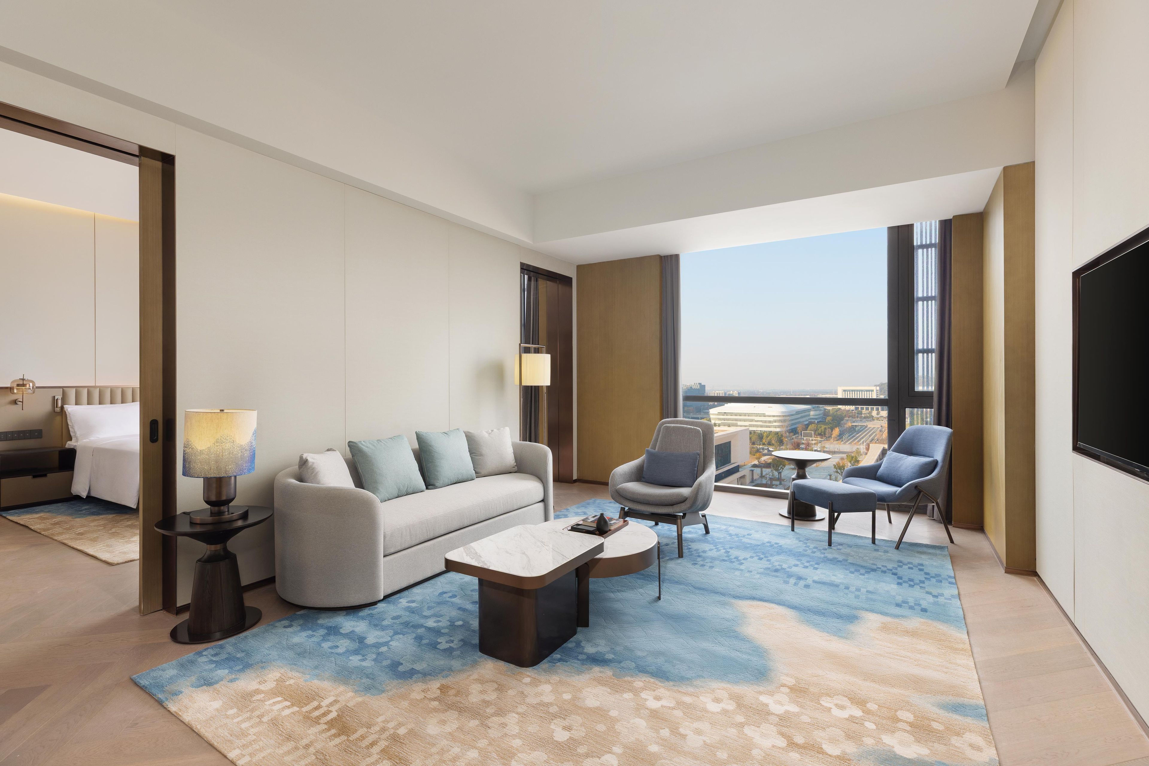 Una habitación de hotel moderna con sofá, sillones, una mesa de centro y vistas a la ciudad a través de amplias ventanas.