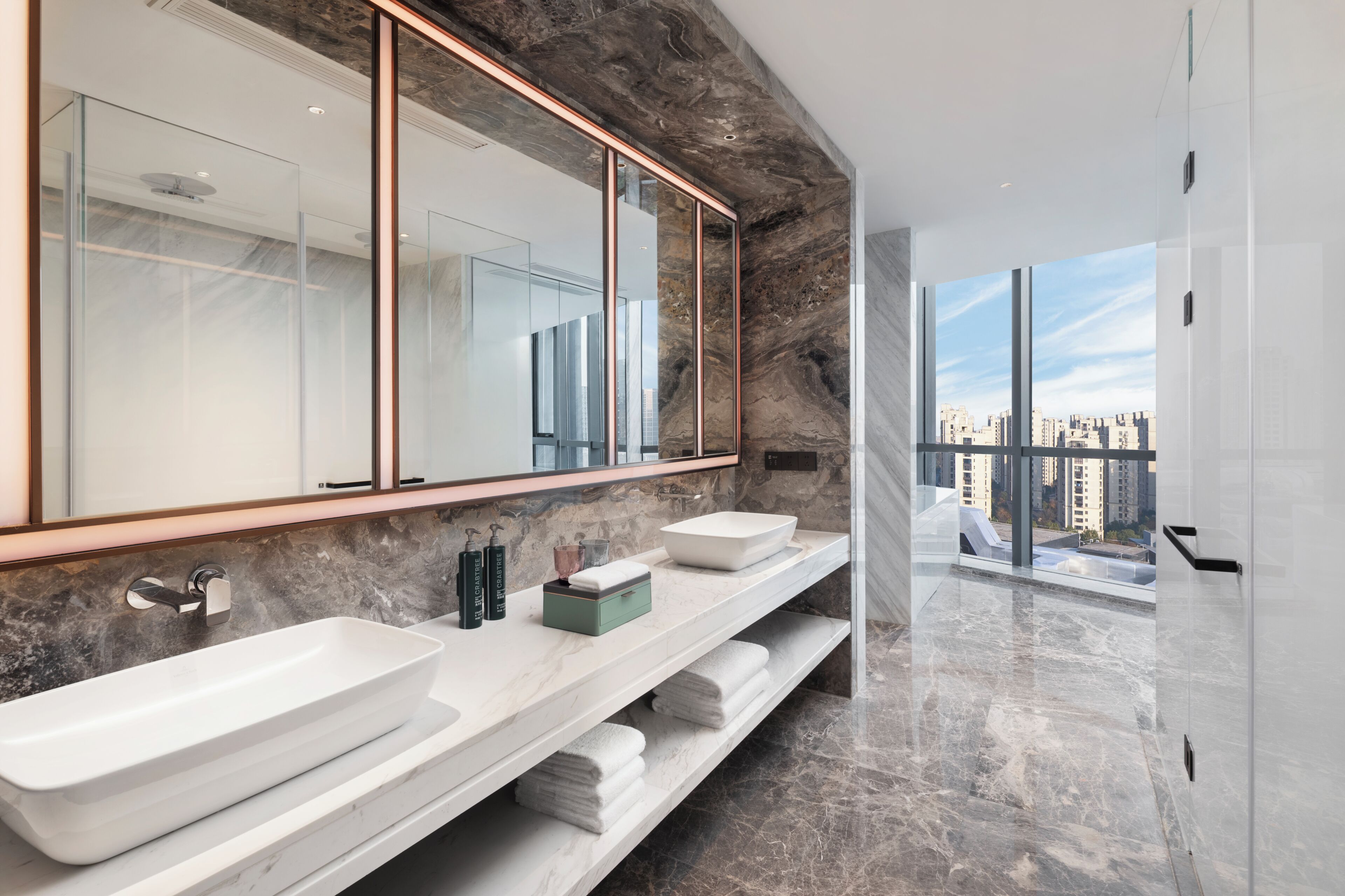 A modern bathroom with a large mirror, two sinks, marble countertops, and a view of the city.