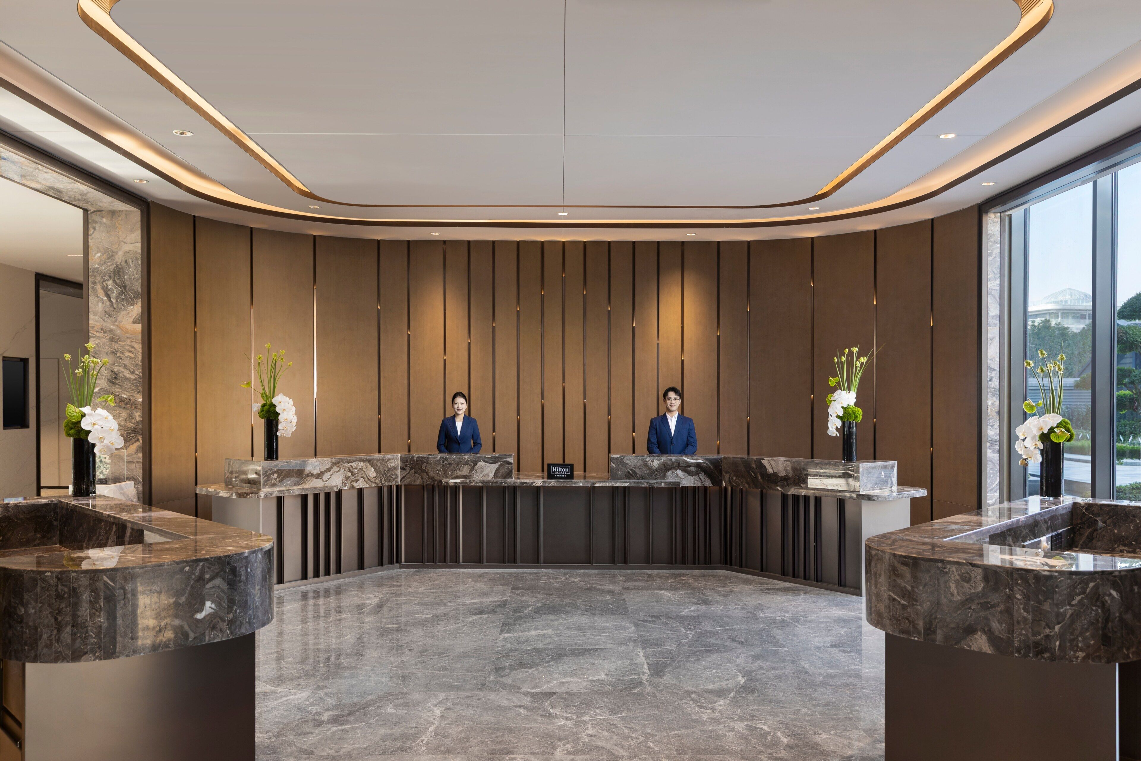 A hotel reception area with a marble reception desk, two receptionists, and a large window providing a view of the outdoors.