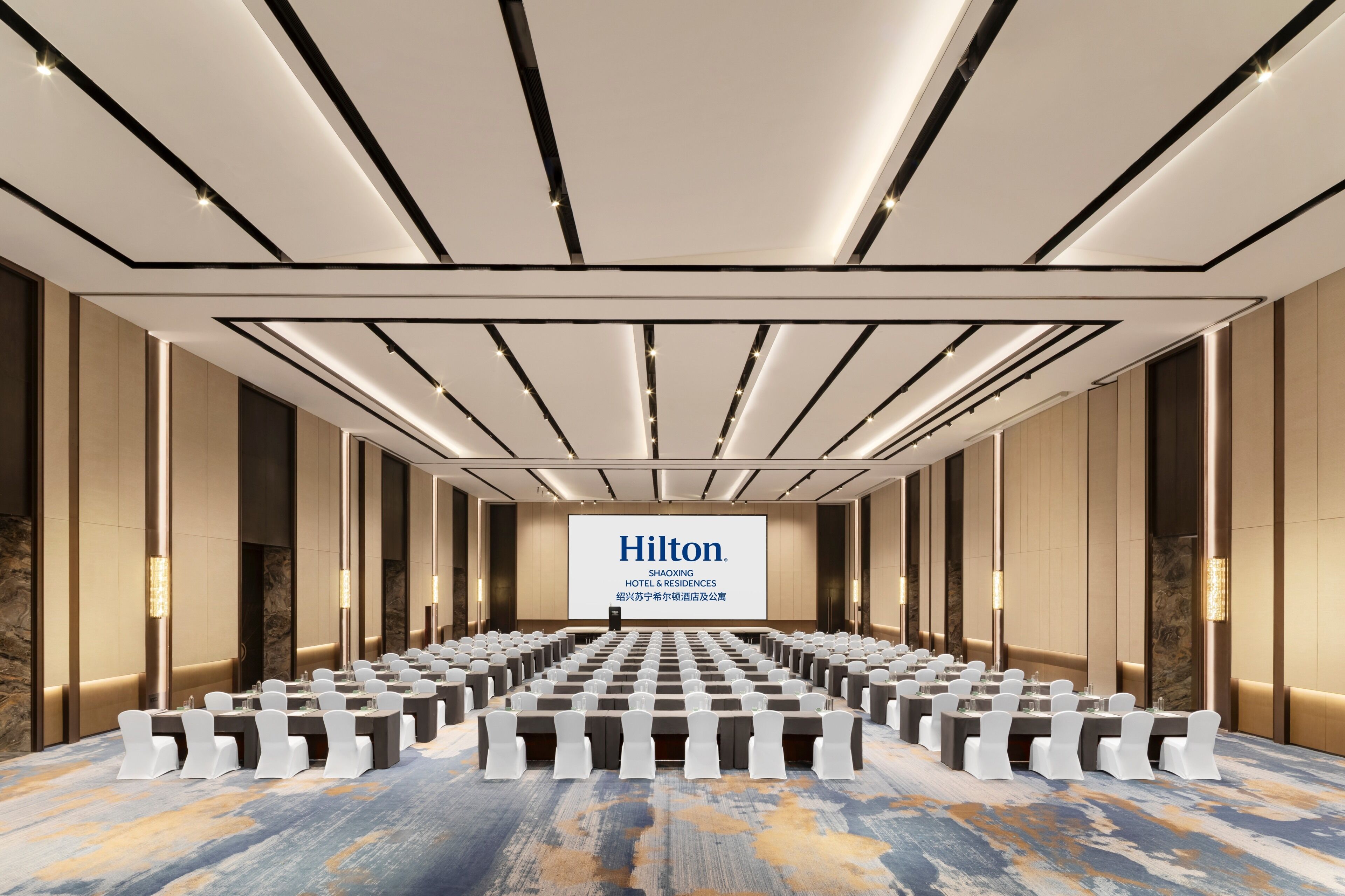 A large conference room with rows of tables and chairs, a large screen displaying 'Hilton,' and a modern ceiling design.