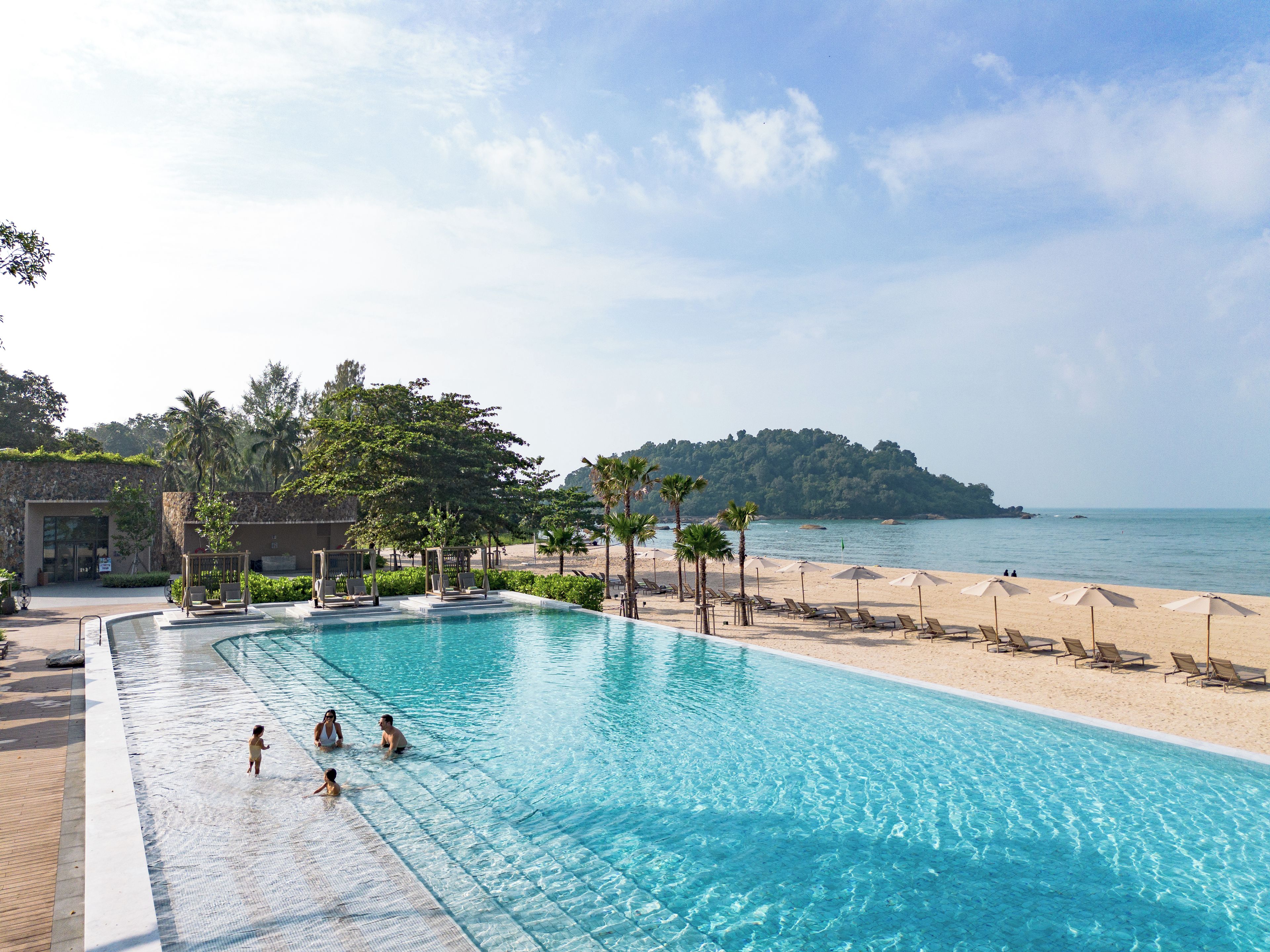 A swimming pool with clear blue water, surrounded by lounge chairs and umbrellas, with a beach and forested hill in the background.
