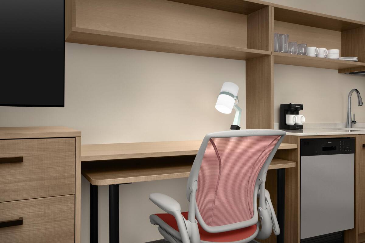 Une chambre d’hôtel moderne équipée d’un bureau en bois, d’une chaise, d’une machine à café, d’un lave-vaisselle et d’une étagère avec des verres et des tasses.