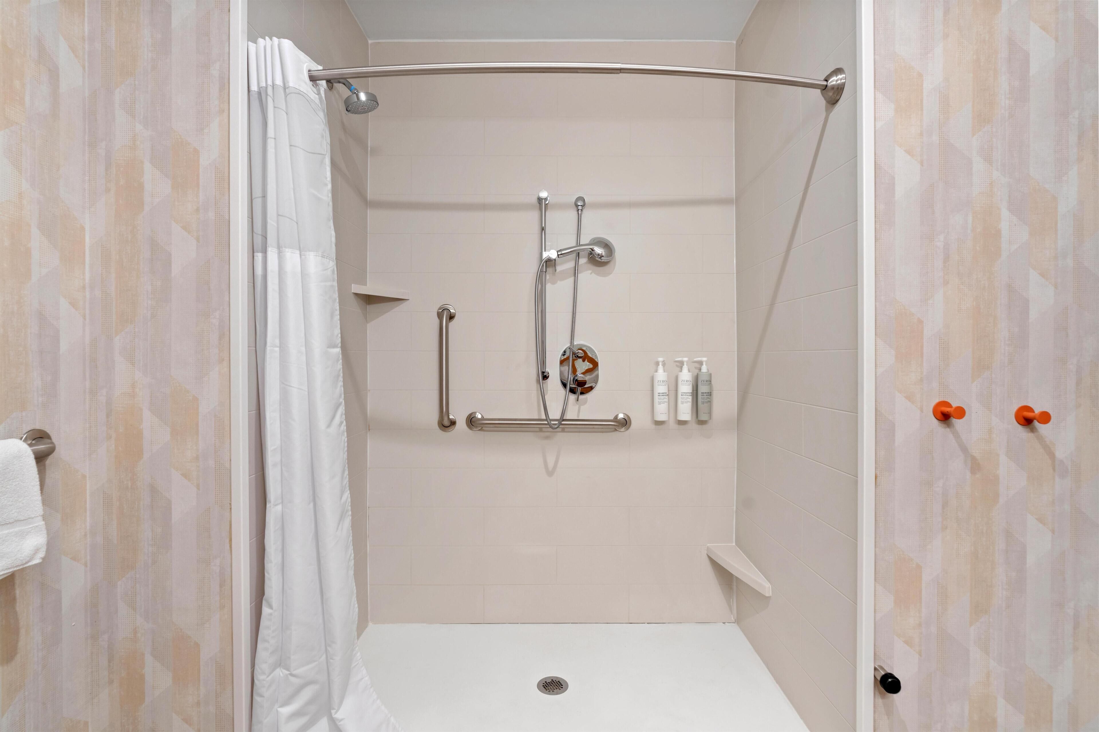 Eine Dusche mit gebogener Duschbrause, einer Handbrause und einem Handduschteil. An der Wand befinden sich Pflegeprodukte und ein Handtuch auf einem Ablagekorb.