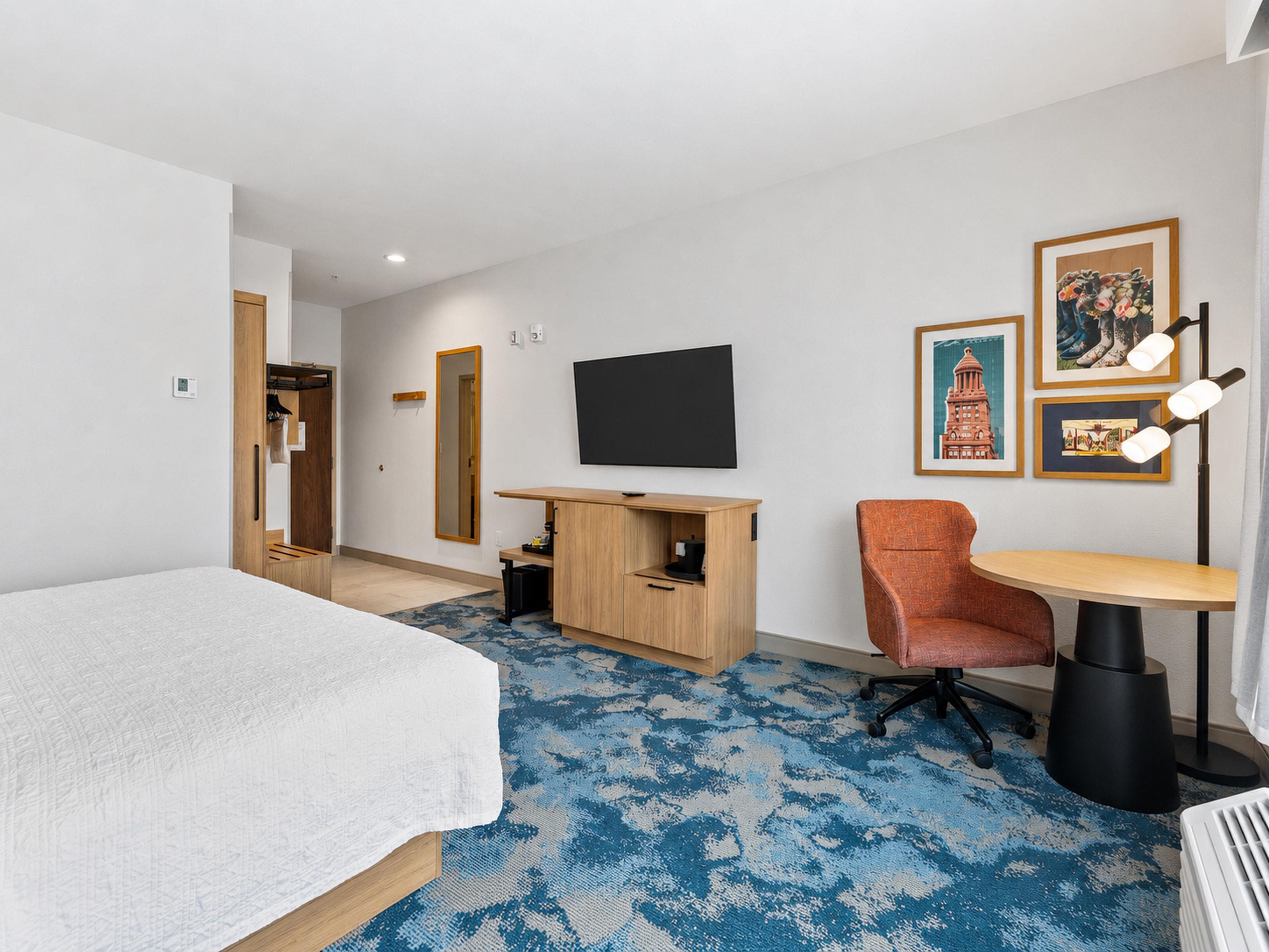 A hotel room with a bed, a desk, a chair, a television, and artwork on the wall.