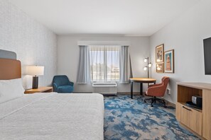 Une chambre d’hôtel avec un lit, un bureau, une chaise, une petite table, une lampe, un radiateur et des tableaux encadrés au mur.