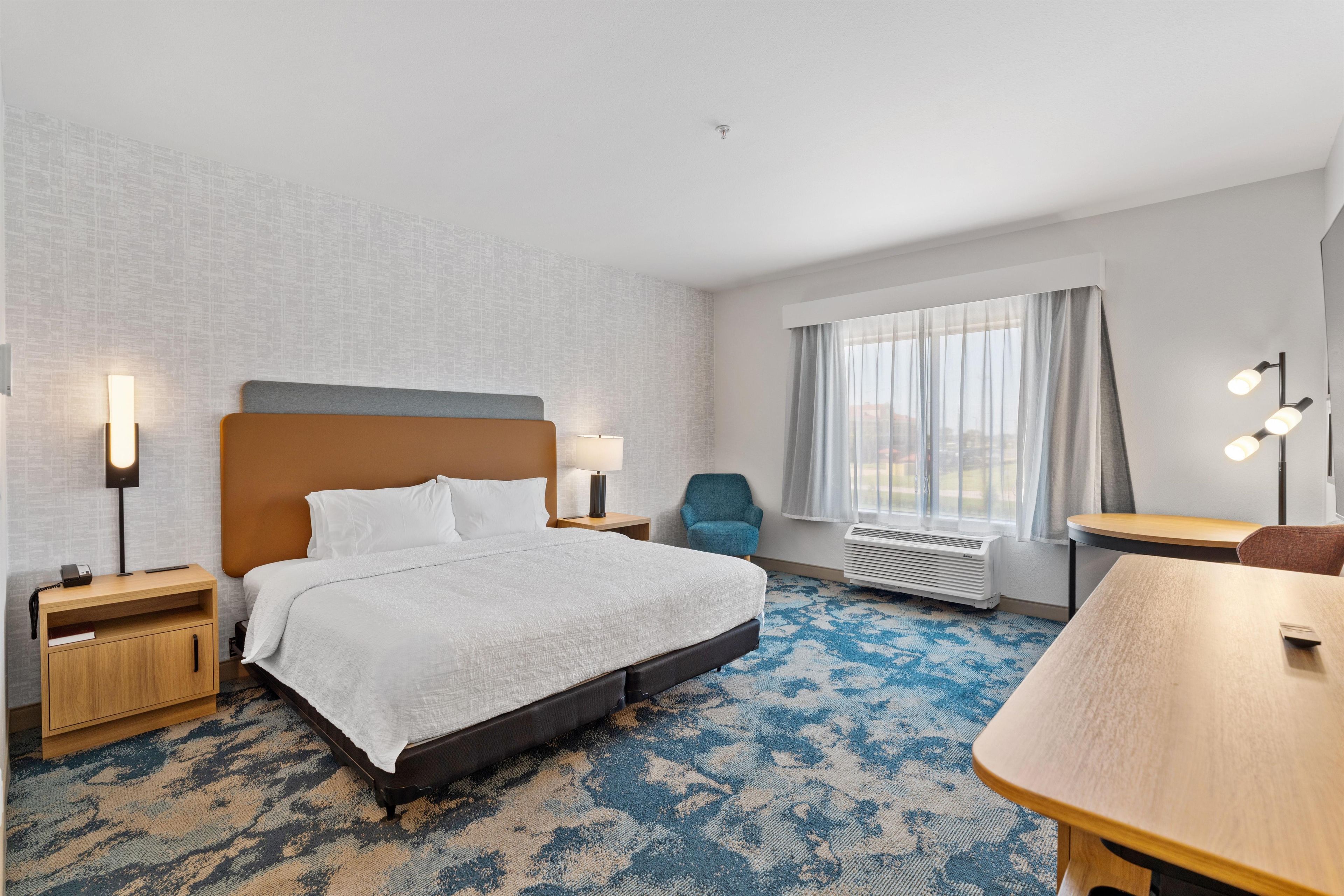 Una habitación de hotel con cama, mesitas de noche, escritorio, silla, ventana con cortinas y unidad de aire acondicionado.