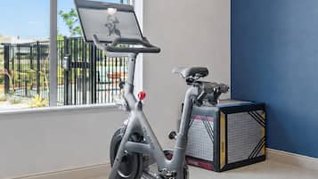 A stationary bike with a screen, a small seat, and pedals, placed in a room with a window and a blue wall.