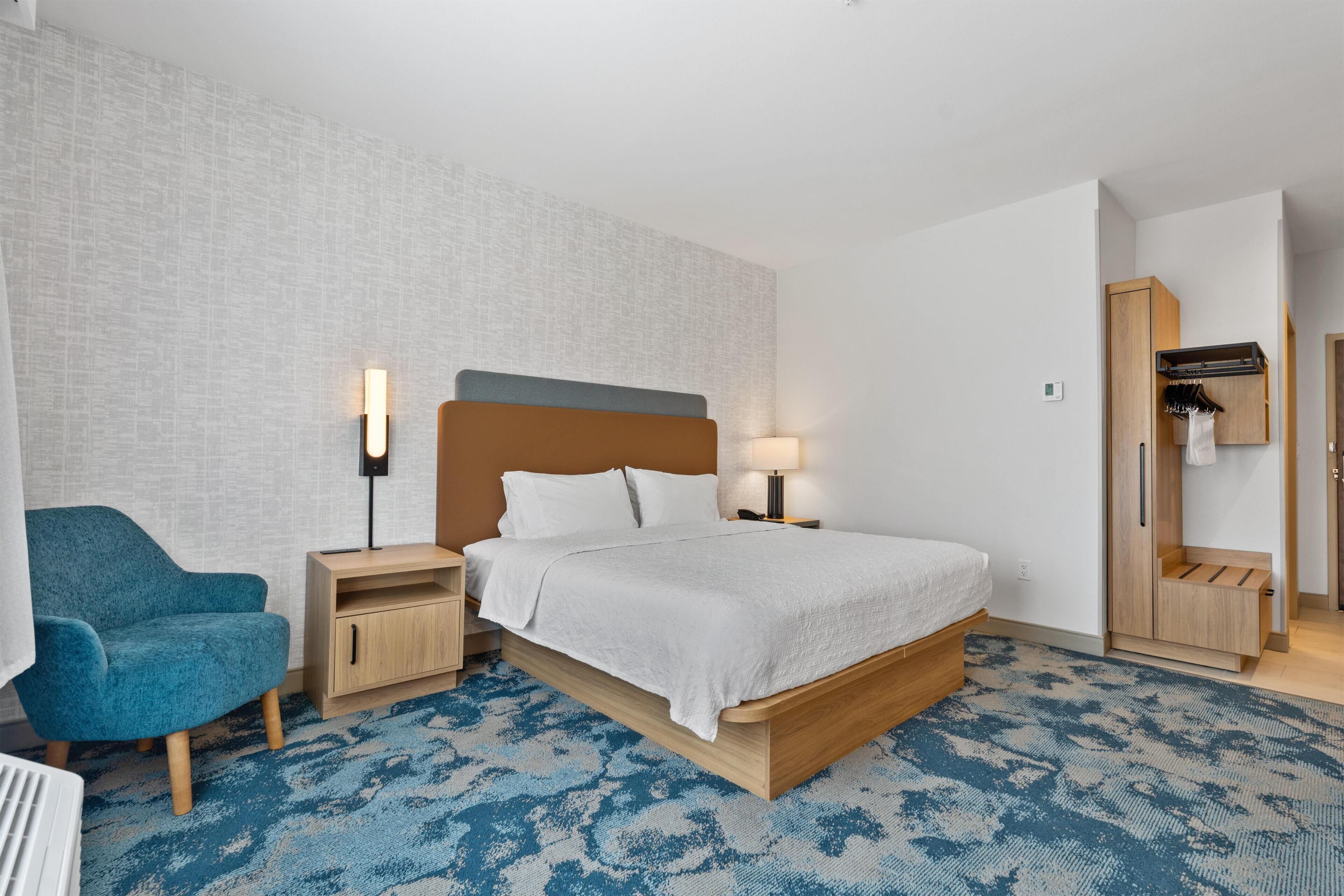 A hotel room with a bed, a chair, a nightstand, a lamp, a wardrobe, and a mirror.