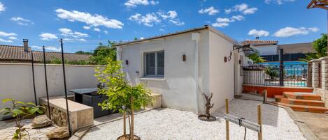 Un petit bâtiment blanc avec une fenêtre, entouré d’une terrasse en gravier agrémentée de plantes en pot et d’une clôture en métal.