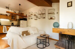 A cozy living room with a white sofa, a small round table, a fireplace, and a kitchen area with wooden cabinets and a hanging light fixture.