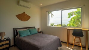 A bedroom with a bed, a nightstand, a desk, and a window with a view of greenery. - Beachfront Ocean-View Apartment in Gated Community (Parrita)