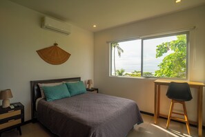 A bedroom with a bed, a nightstand, a desk, and a window with a view of greenery. - Beachfront Ocean-View Apartment in Gated Community (Parrita)