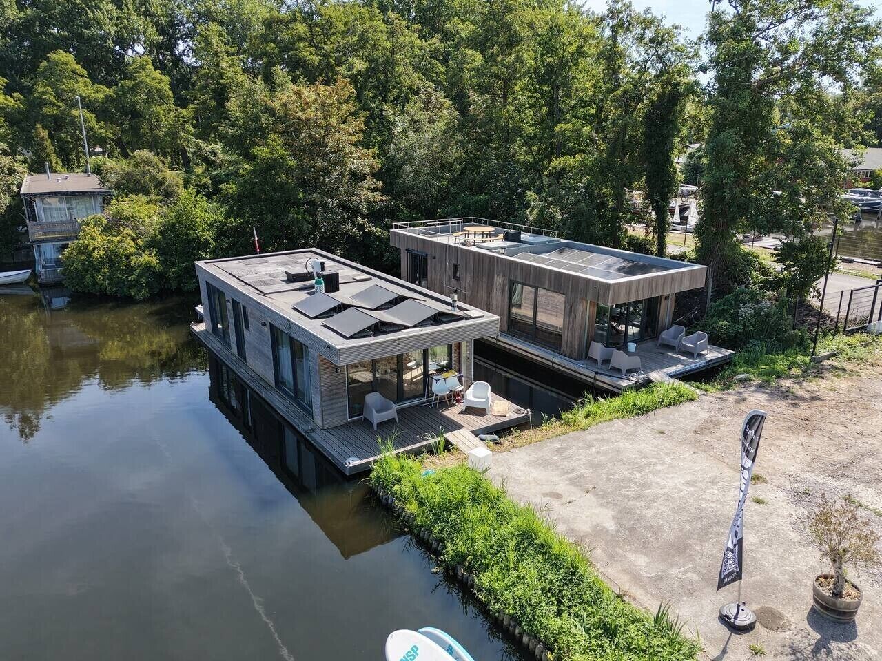 Ein modernes Hausboot mit Deck und Außenbereich, an einem mit Bäumen umgebenen Gewässer angedockt.
