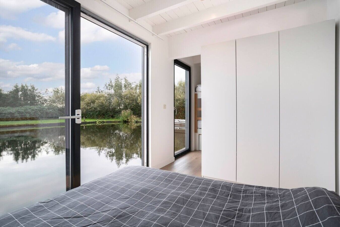 Een slaapkamer met een groot raam, een bed met een beddeken met een patroon, en uitzicht op een meer en bomen.