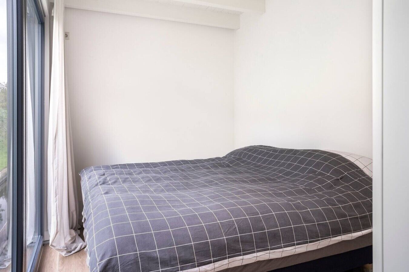 Una cama individual con una colcha a cuadros en una habitación con paredes blancas y una ventana.
