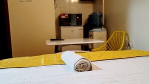 Quarto de hotel com cama, televisão fixada na parede exibindo um canal, micro-ondas, mesa pequena e uma cadeira.