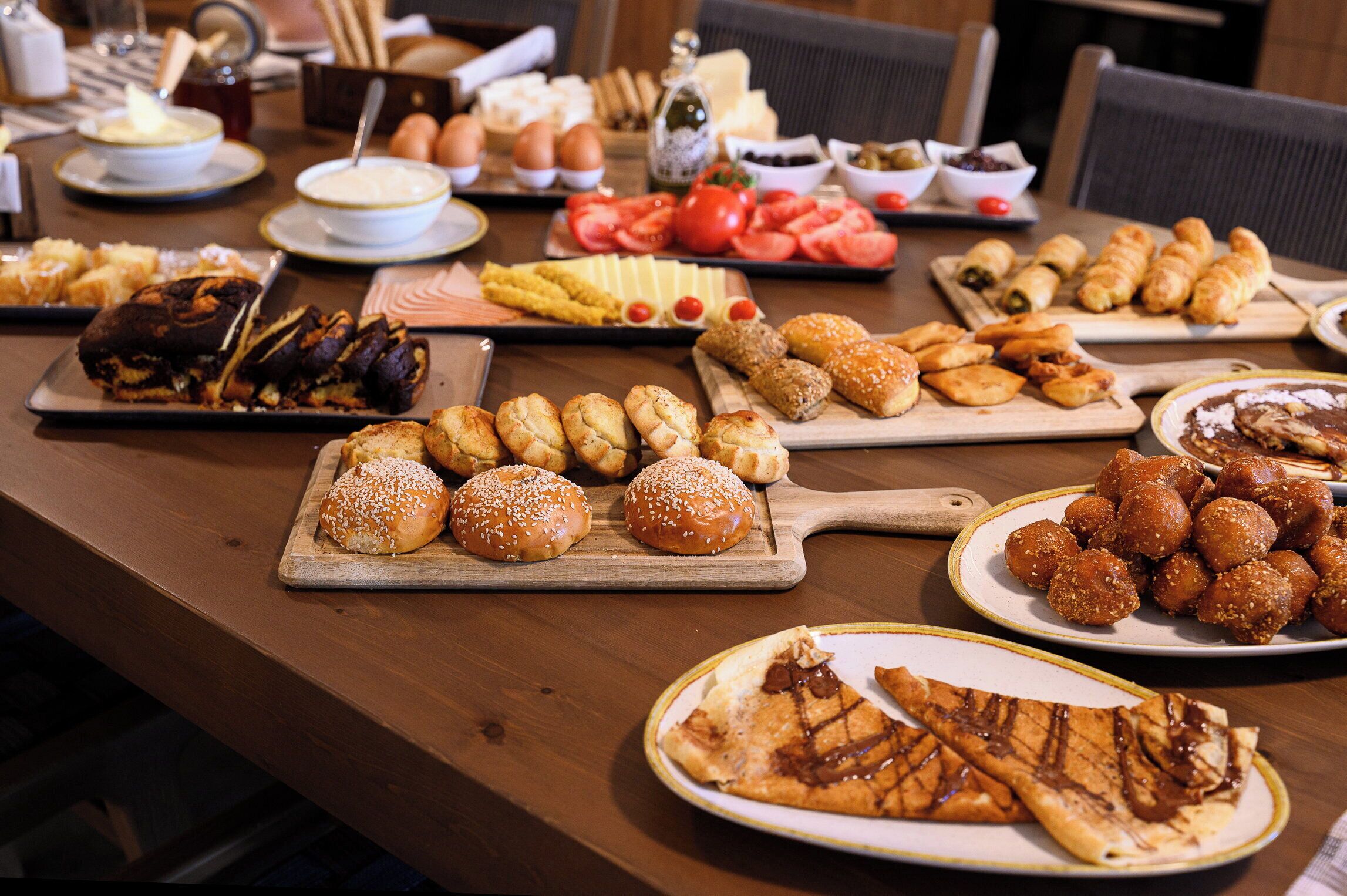 A variety of food items on a wooden table, including pastries, bread, and sliced tomatoes.