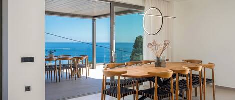 A modern dining area with a wooden table and chairs, large windows with a view of the sea, and a hanging light fixture.