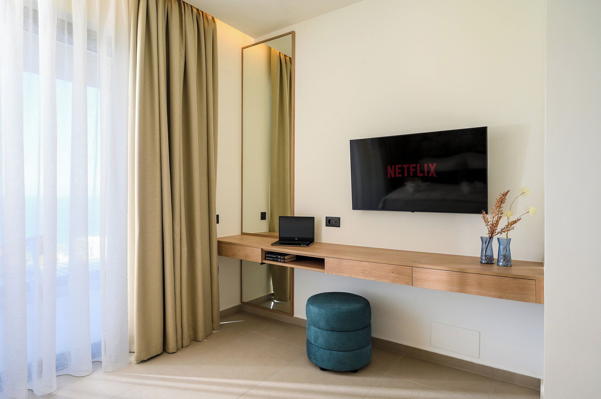 A modern hotel room with a wooden desk, a laptop, a TV mounted on the wall, a mirror, and a blue ottoman.