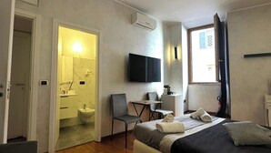 A modern hotel room with a bed, a desk, a chair, a TV, and a bathroom visible through an open door. - The rooms in the People's Square in Rome (Roma)
