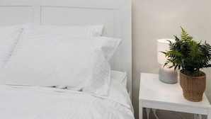 A bedroom with a white bed, a white nightstand with a lamp and a plant, and a wall-mounted picture of a palm tree.