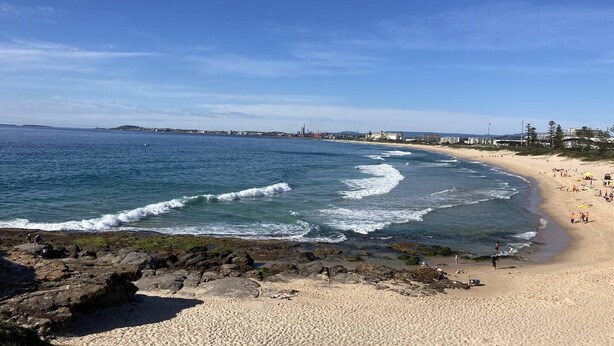 A beach with clear blue water, sandy shore, and buildings in the background.