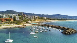 A marina with numerous sailboats docked, a rocky breakwater, and a backdrop of residential buildings and a mountain.