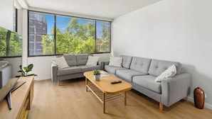 A modern living room with a grey sofa, a wooden coffee table, and a large window offering a view of trees and buildings.