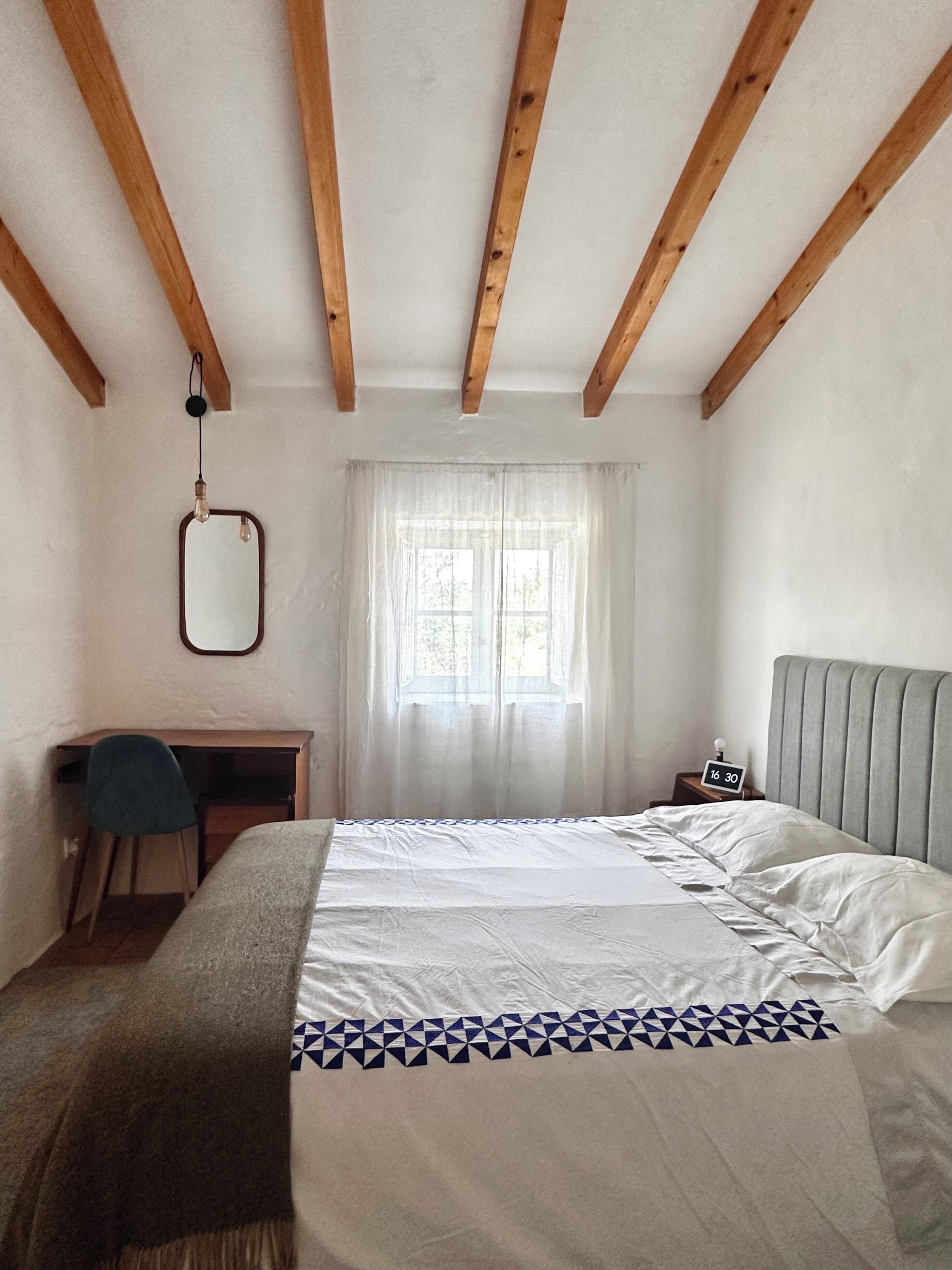 A bedroom with a bed, a desk, a chair, a nightstand, and a window with curtains.
