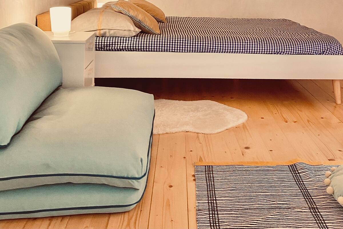A small, cozy room with a sloped ceiling, a bed with a checkered bedspread, a bedside table with a lamp, and a plush armchair.