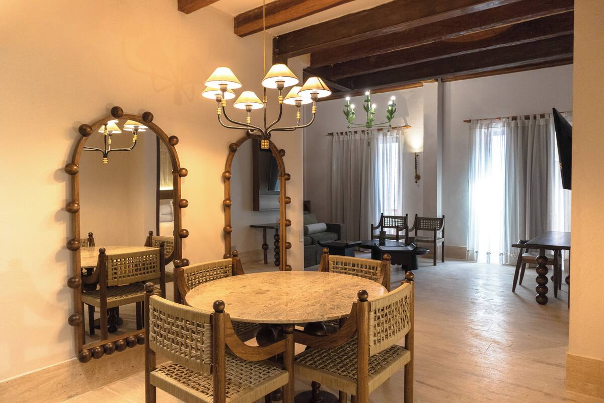 Una zona de menjador amb una taula rodona, cadires de fusta i un penjall. Hi ha dos grans miralls a les parets, i es veu una altra habitació amb un sofà i cortines.