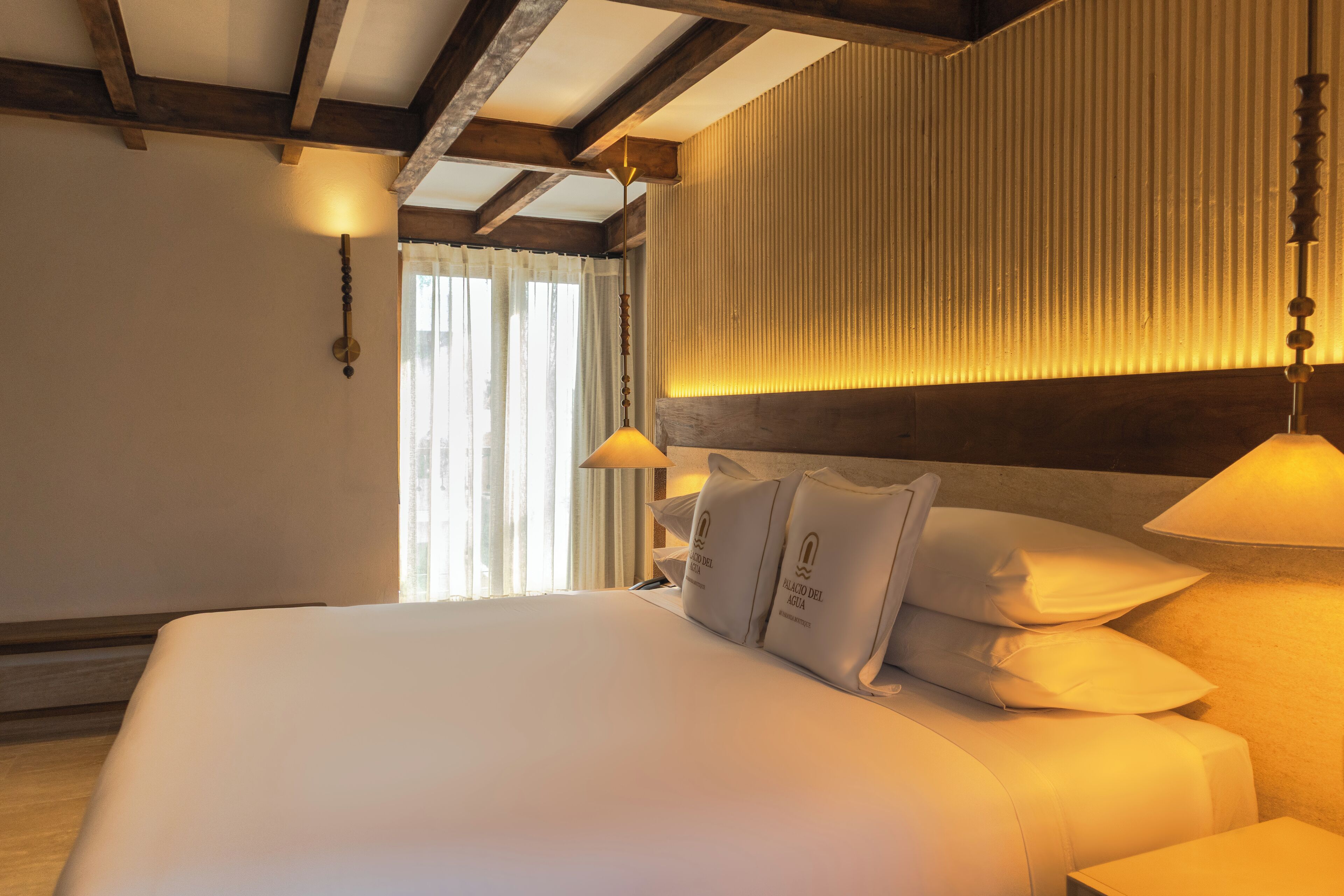 A hotel room with a large bed, a wooden ceiling, a wall-mounted light, and a window with curtains.