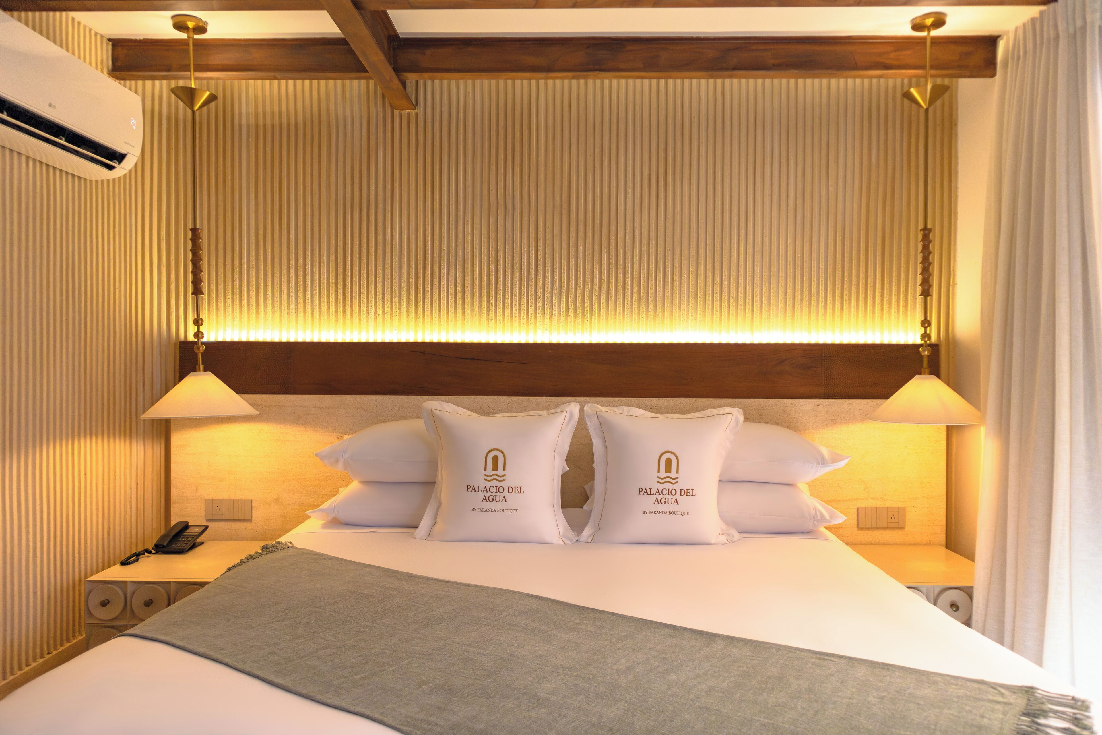 A hotel room with a bed, two bedside lamps, a nightstand, a telephone, and a wall-mounted air conditioner.