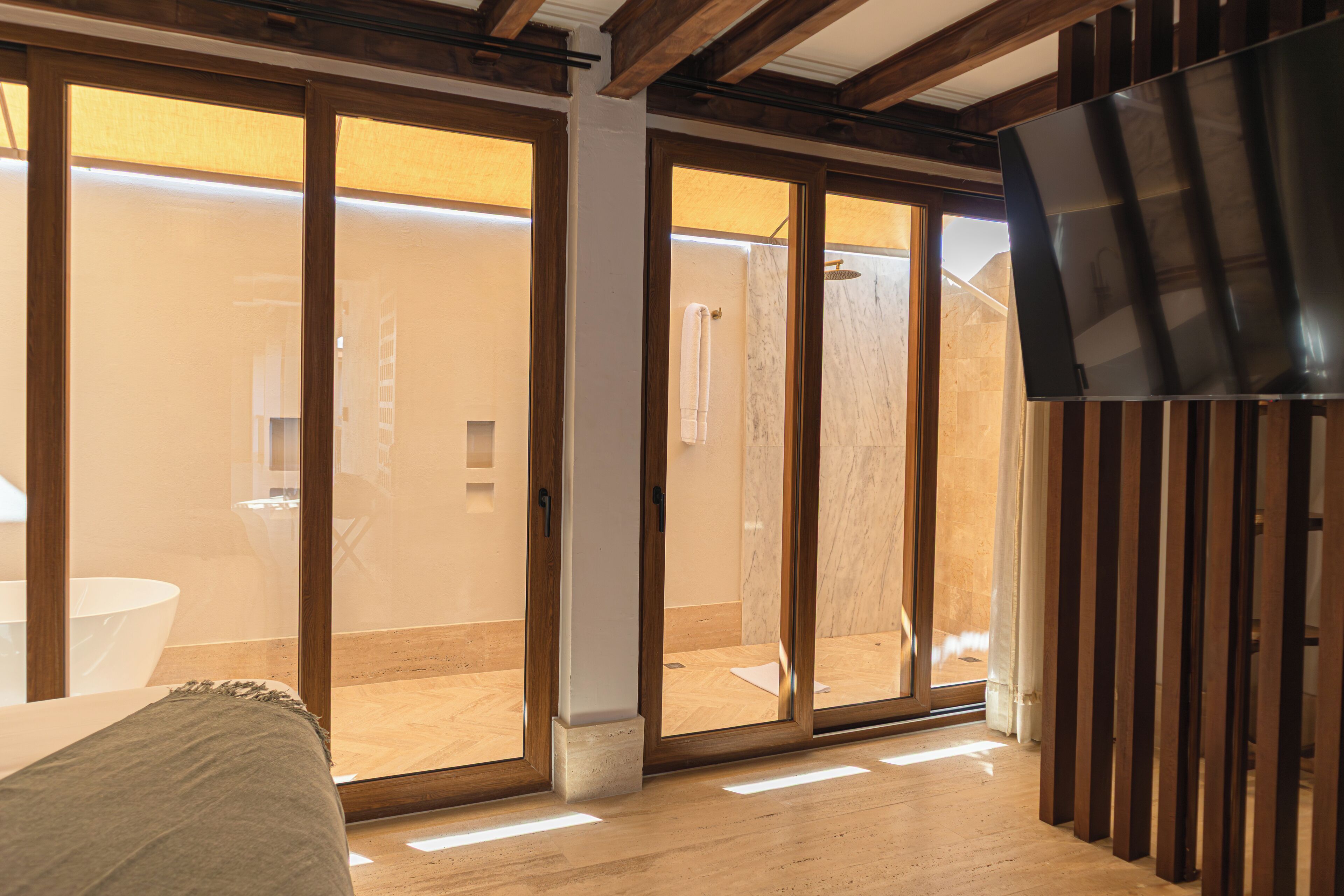 A modern hotel room with a large sliding glass door leading to a bathroom, a wooden ceiling, and a flat-screen TV mounted on the wall.