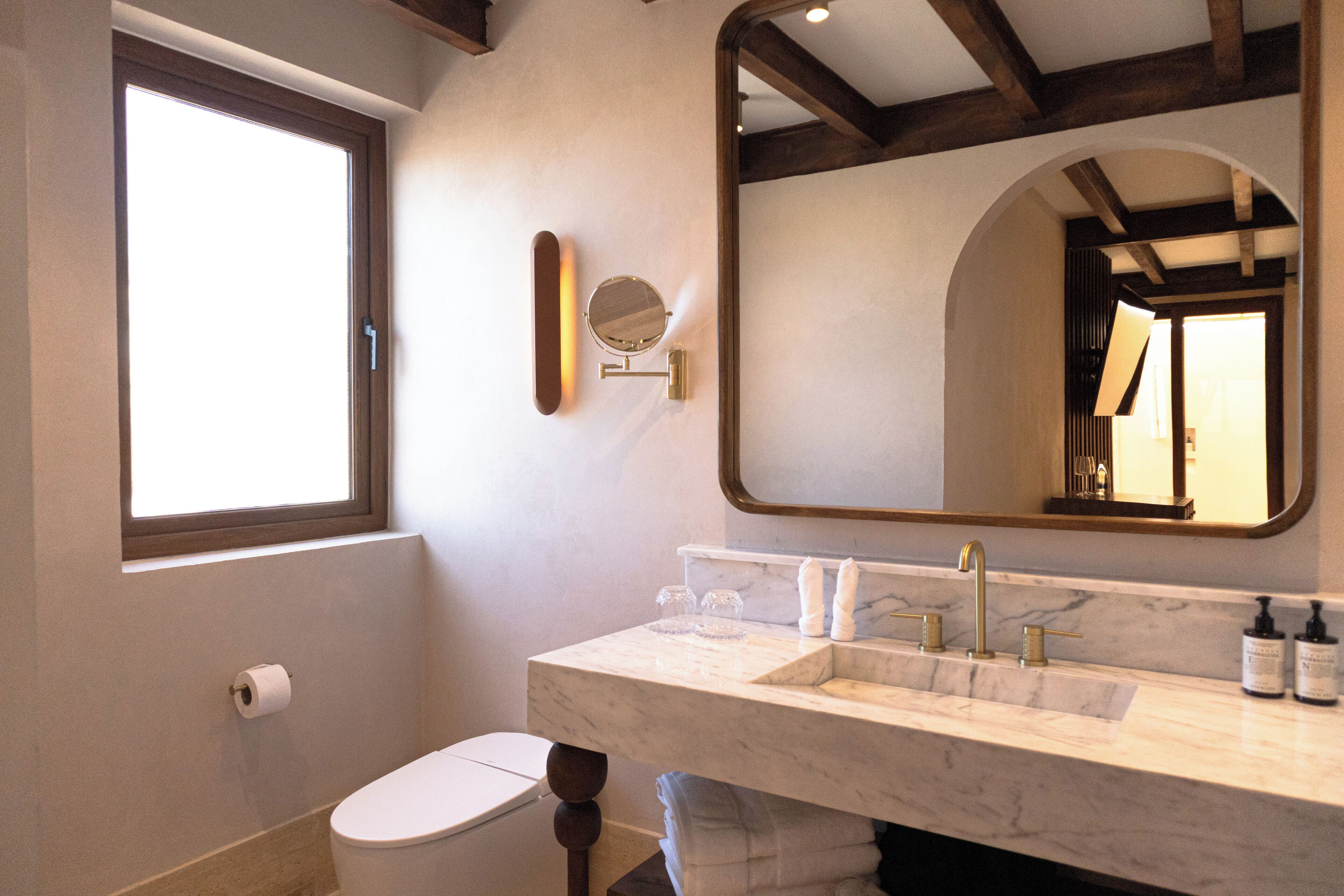A bathroom with a toilet, a sink with a marble countertop, a large mirror, and a window.
