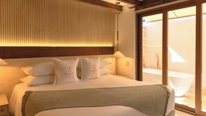 Habitación de hotel con una cama grande, techo de madera, puerta corredera de vidrio hacia el baño y una lámpara de pared.