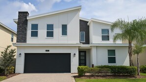 A two-story modern house with a black garage door, a stone chimney, and a well-maintained lawn. - ChampionsGate Resort 8BR Home w/ Pool, Hot Tub, Theater, Game Room  & Much More! (Kissimmee)