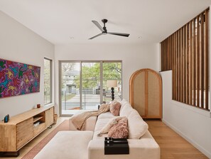 A modern living room with a sofa, a ceiling fan, a wooden cabinet, and a large window. - Modern East Austin Pool Home - Sleeps 8 - 8 Min DT (Austin)