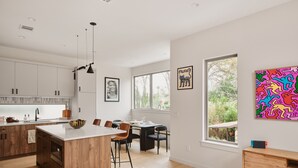 Eine moderne Küche mit Holzschränken, einer zentralen Kochinsel und Pendelleuchten. Es gibt einen Essbereich mit Tisch und Stühlen sowie ein großes Fenster mit Blick auf Grünflächen.