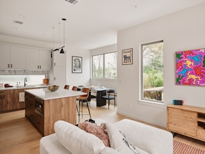 A modern kitchen with wooden cabinets, a central island, and pendant lighting. There is a dining area with a table and chairs, and a large window offering a view of greenery. - Modern East Austin Pool Home - Sleeps 8 - 8 Min DT (Austin)